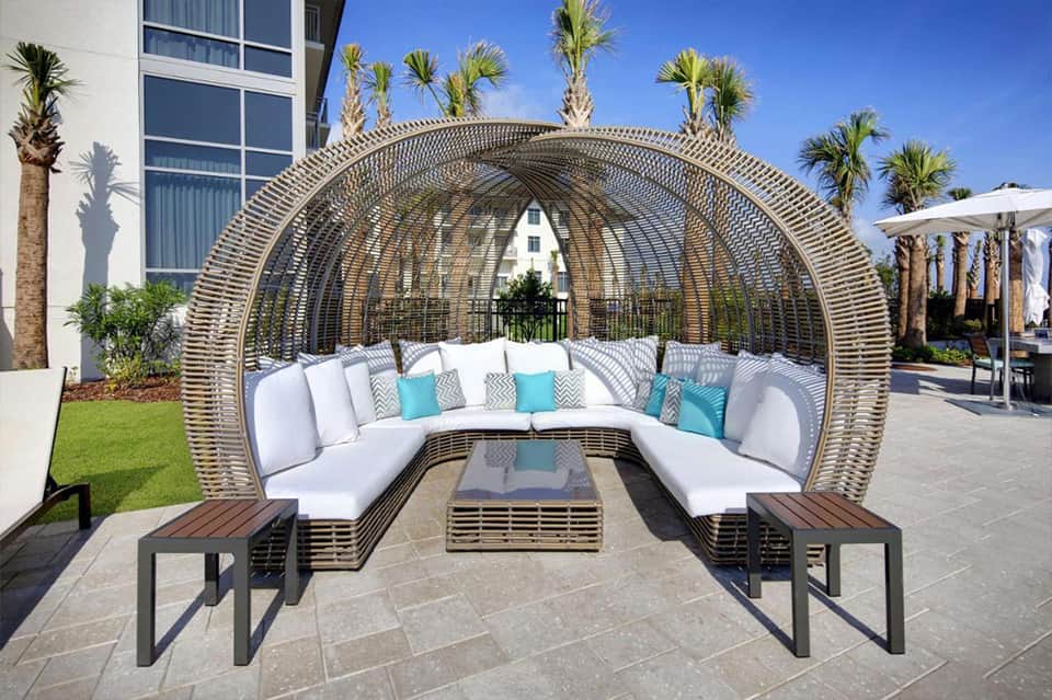 Modern woven lounge chair with white cushions and turquoise pillows on palm-lined patio