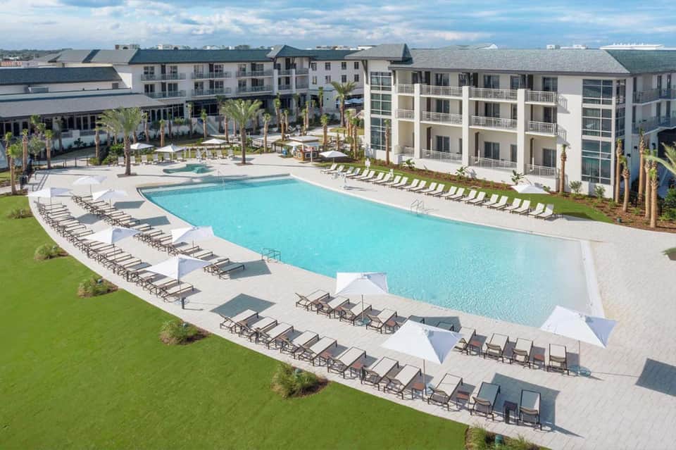 Modern resort with curved pool, lounge chairs, palm trees, and multi-story apartment buildings under cloudy sky
