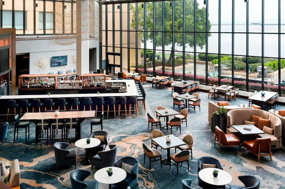 Modern hotel lobby with high ceilings, bar seating, lounge areas, and floor-to-ceiling windows overlooking water