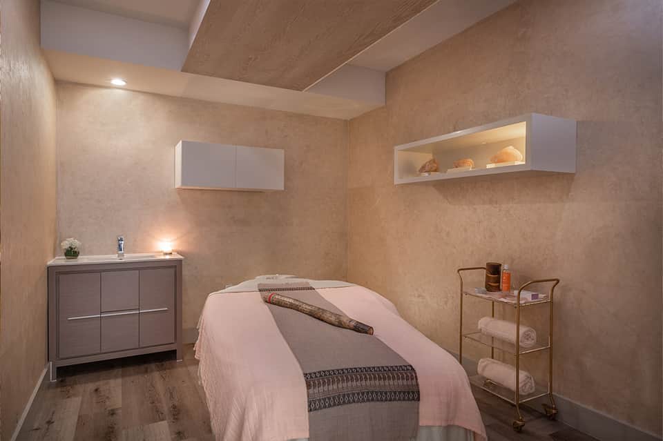 Spa treatment room with warm beige walls, massage bed, wooden storage cabinet, and shelving with folded towels