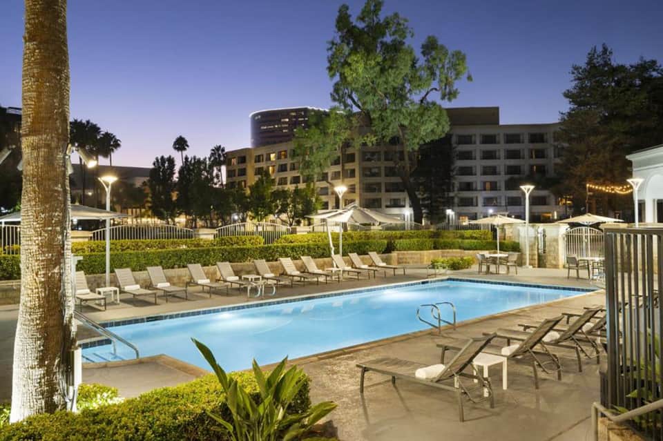Resort swimming pool with lounge chairs at dusk, surrounded by palm trees and modern hotel buildings