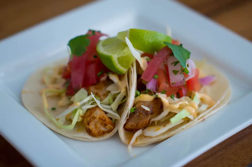 Two tacos with shredded chicken, lime, red onion, cilantro, and cabbage on a white plate