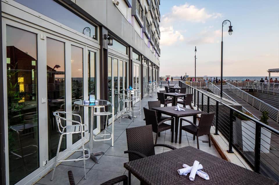 Waterfront terrace with black dining tables, white chairs, and views of the bay and promenade