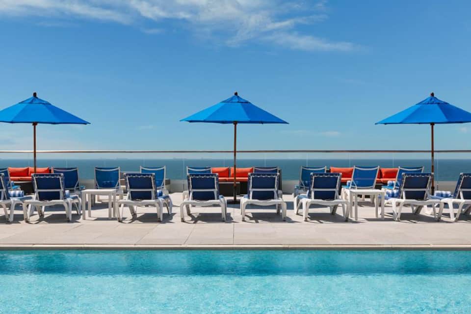 Oceanfront pool deck with blue umbrellas, white lounge chairs, and panoramic ocean view on clear day