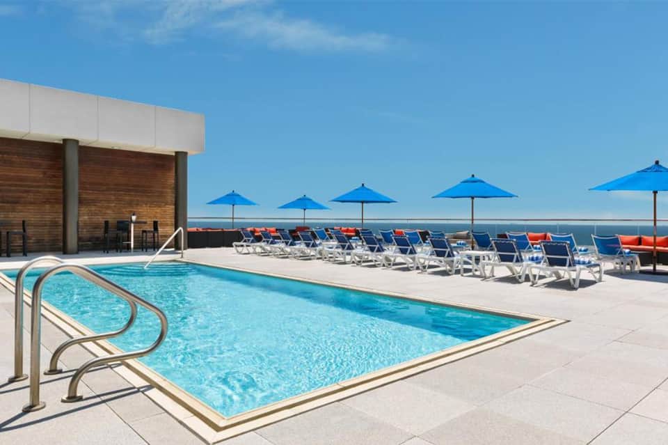 Resort pool deck with blue umbrellas, white lounge chairs, and ocean view on clear sunny day