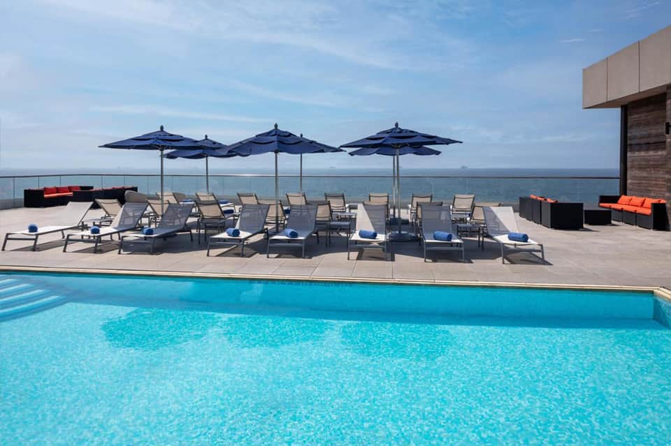 Modern resort pool with blue umbrellas, lounge chairs, and ocean view with waterfront landscape