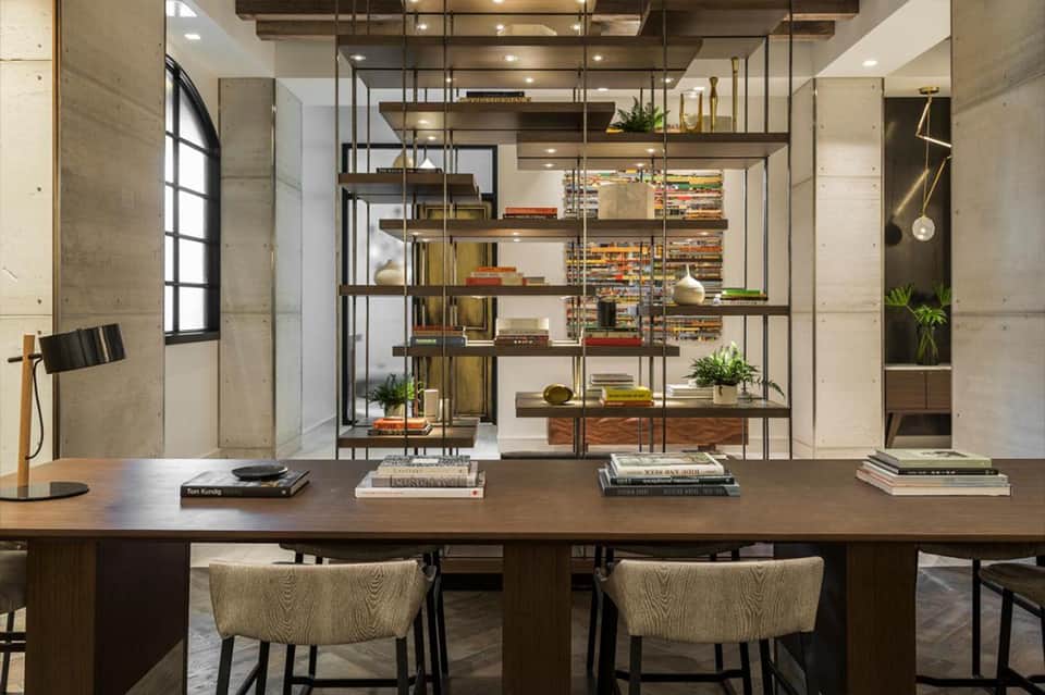Modern office workspace with wooden desk, woven chairs, and floor-to-ceiling metal shelving displaying books and decorative items