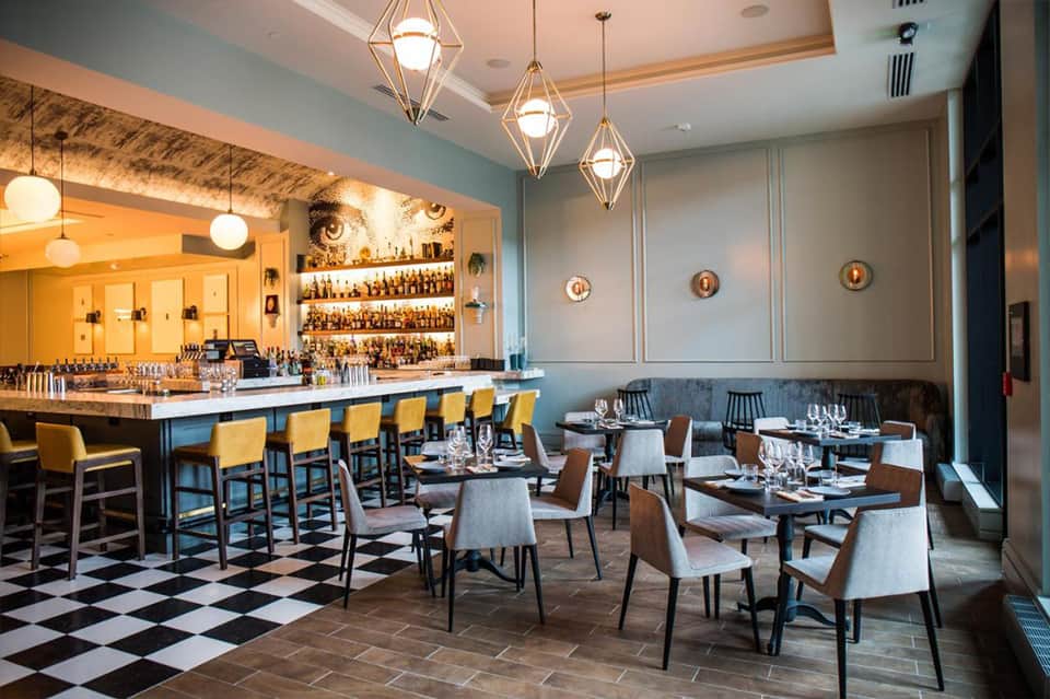 Modern restaurant interior with blue walls, bar seating with yellow chairs, pendant lights, and dining tables