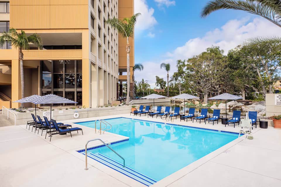Resort outdoor pool with blue water, loungers, umbrellas, and multi-story building with palm trees