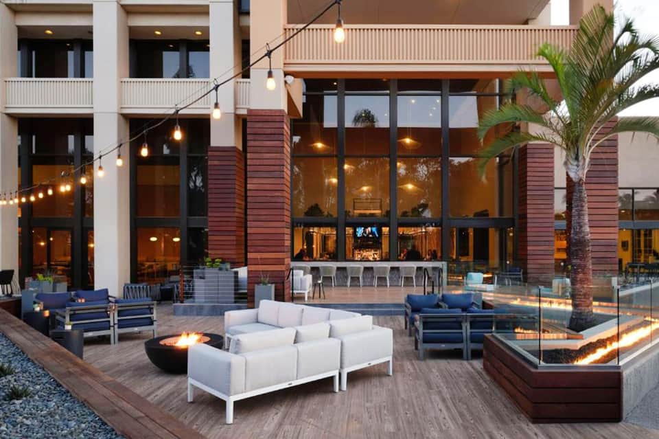 Contemporary courtyard with white seating, fire pit, palm trees, and modern building with warm lighting at dusk