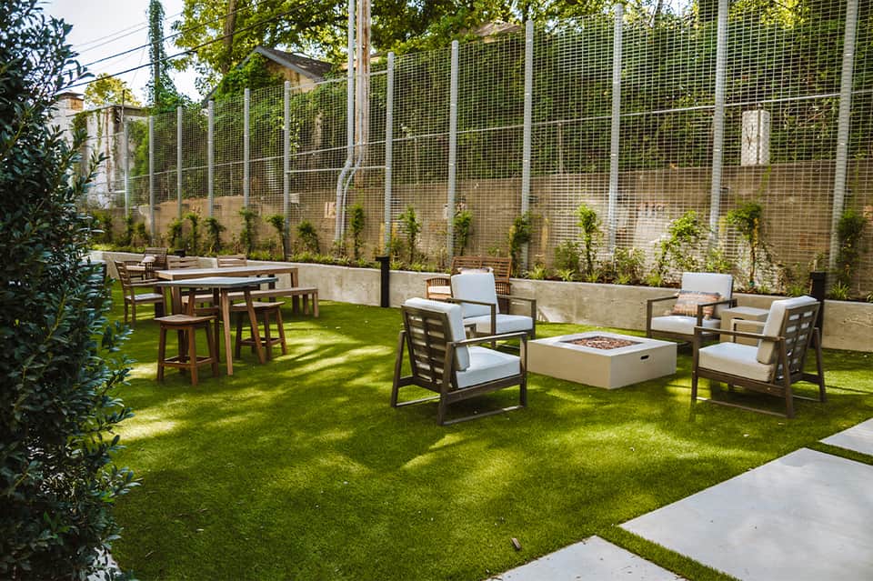 Modern backyard with green lawn, wooden dining table, lounge chairs, fire pit, and metal fence screening