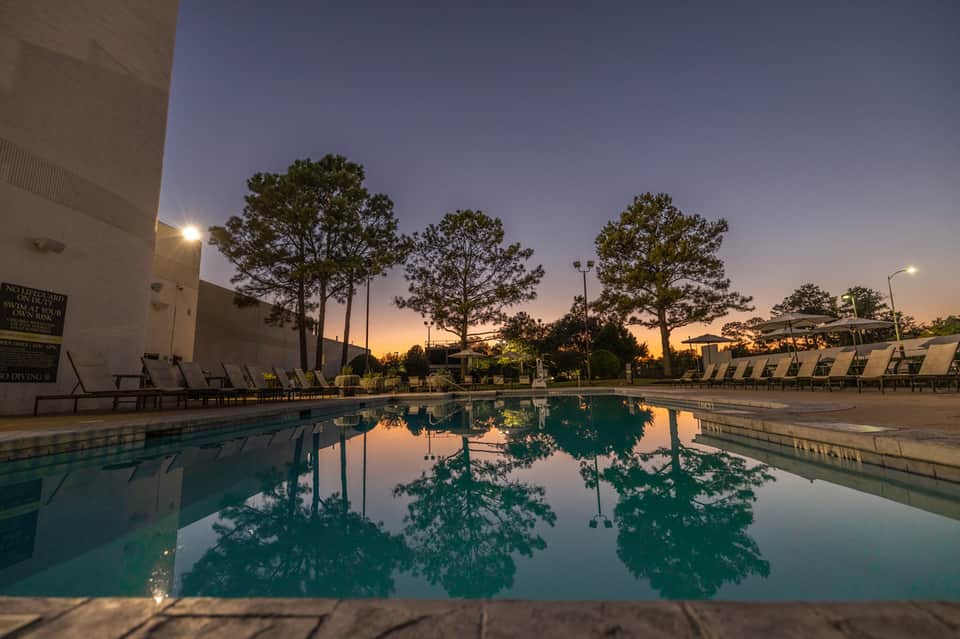 Resort swimming pool at dusk with lounge chairs and tall pine trees reflected in calm water