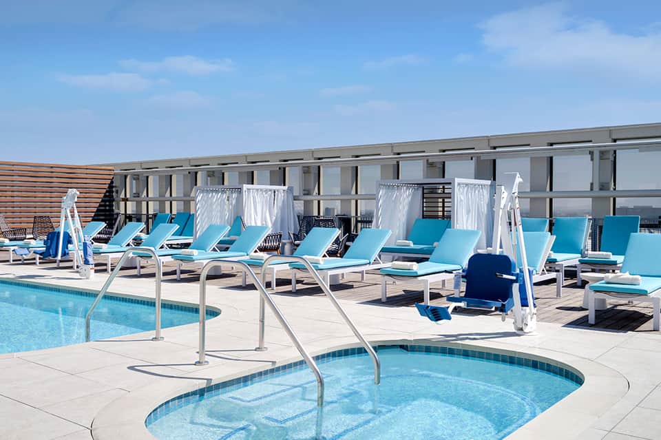 Rooftop pool deck with bright blue lounge chairs, white cabanas, and modern building with glass railings