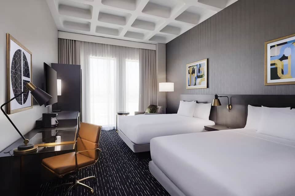 Modern hotel room with dark gray accent wall, two beds, workspace with monitor, coffered white ceiling, and contemporary furnishings
