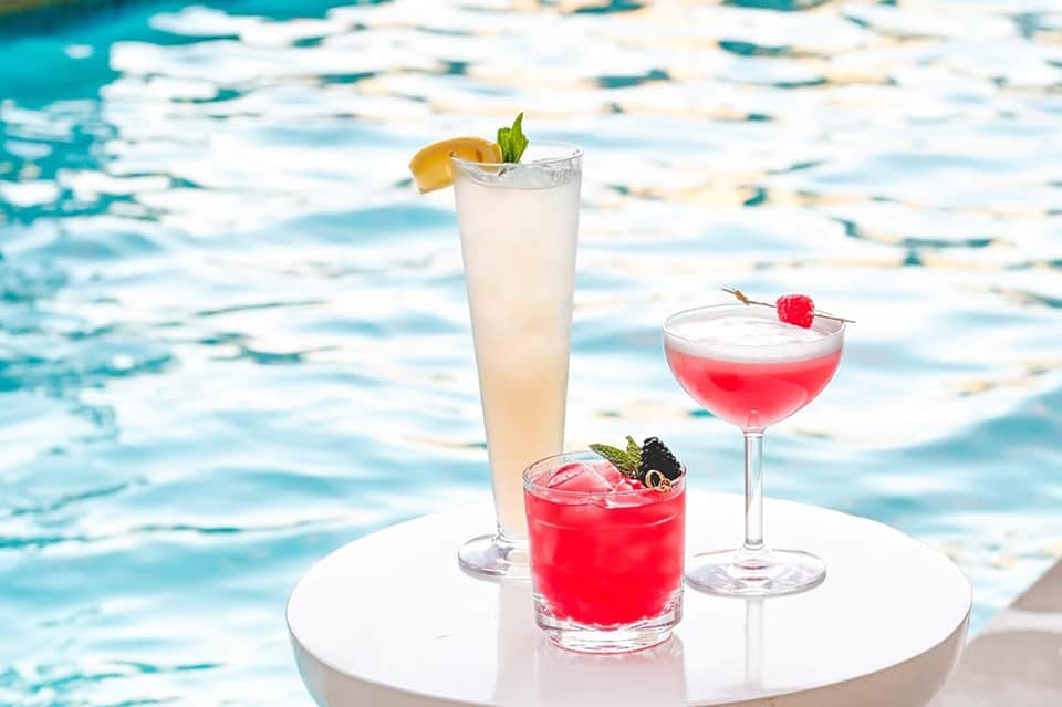 Three colorful cocktails on white table by sparkling pool with tropical garnishes