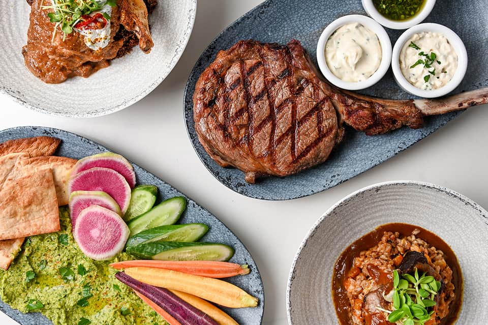Upscale restaurant dishes featuring grilled steak, green sauce, fresh vegetables, and creamy dips on elegant plating