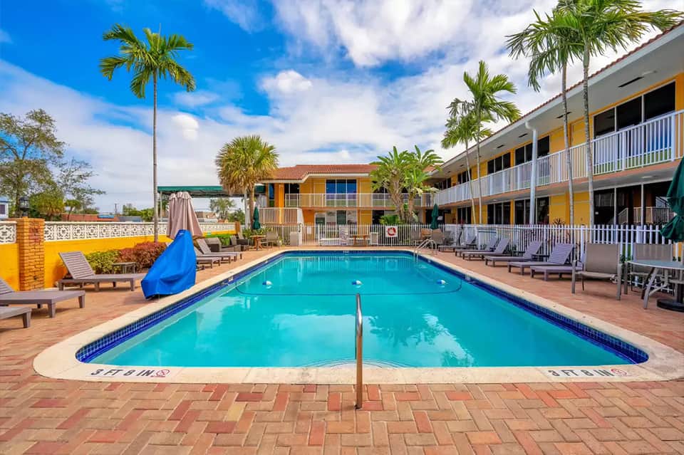 Resort pool surrounded by palm trees, loungers, and yellow and blue-trimmed buildings under clear sky