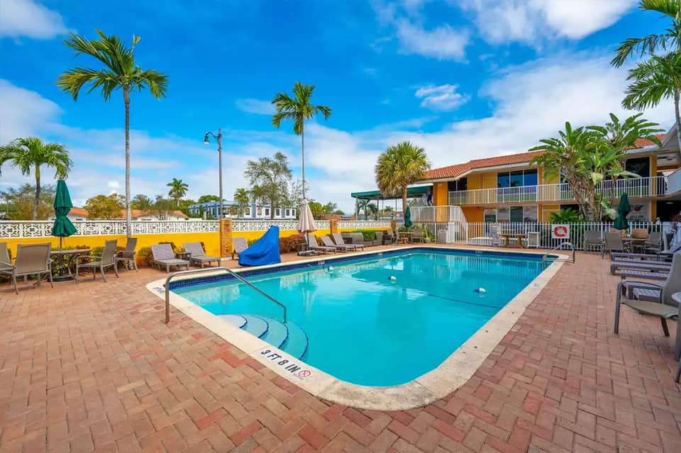 Resort pool surrounded by palm trees with yellow loungers and colorful umbrellas, brick patio, ocean view in background
