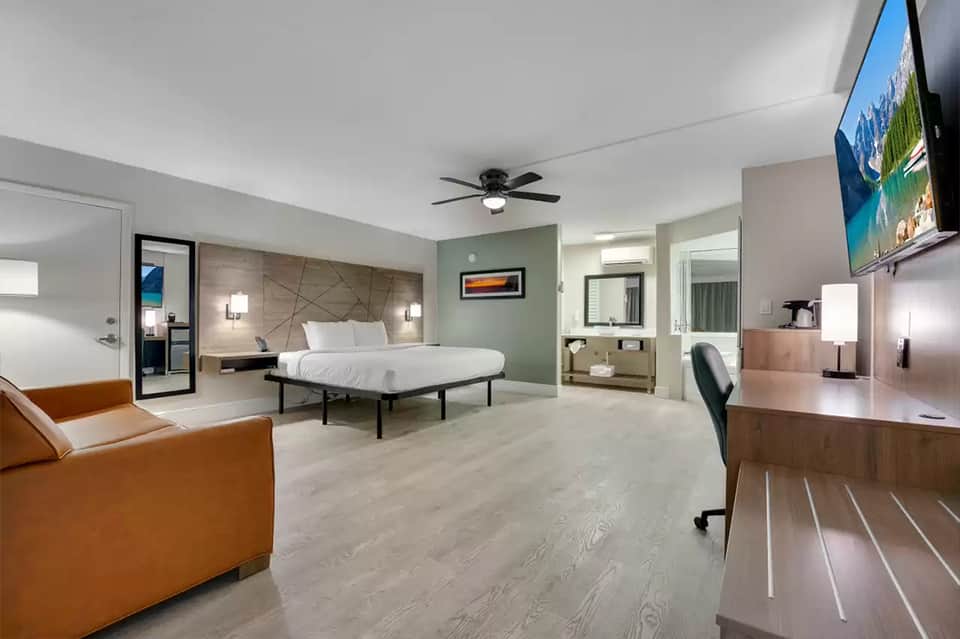 Spacious hotel suite with king bed, living area with tan leather chair, desk workspace, and ensuite bathroom