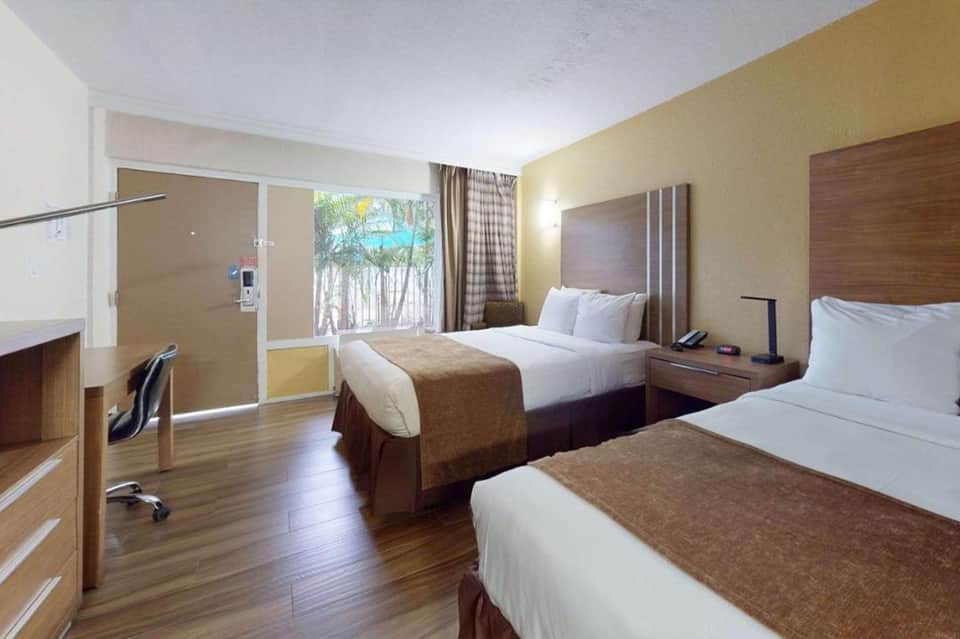 Hotel room with two queen beds, wooden furniture, yellow walls, and window overlooking palm trees