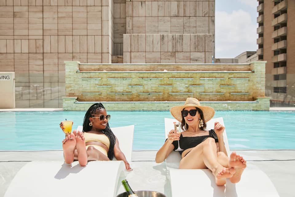 Two women lounging poolside with cocktails, enjoying sun and sparkling pool water