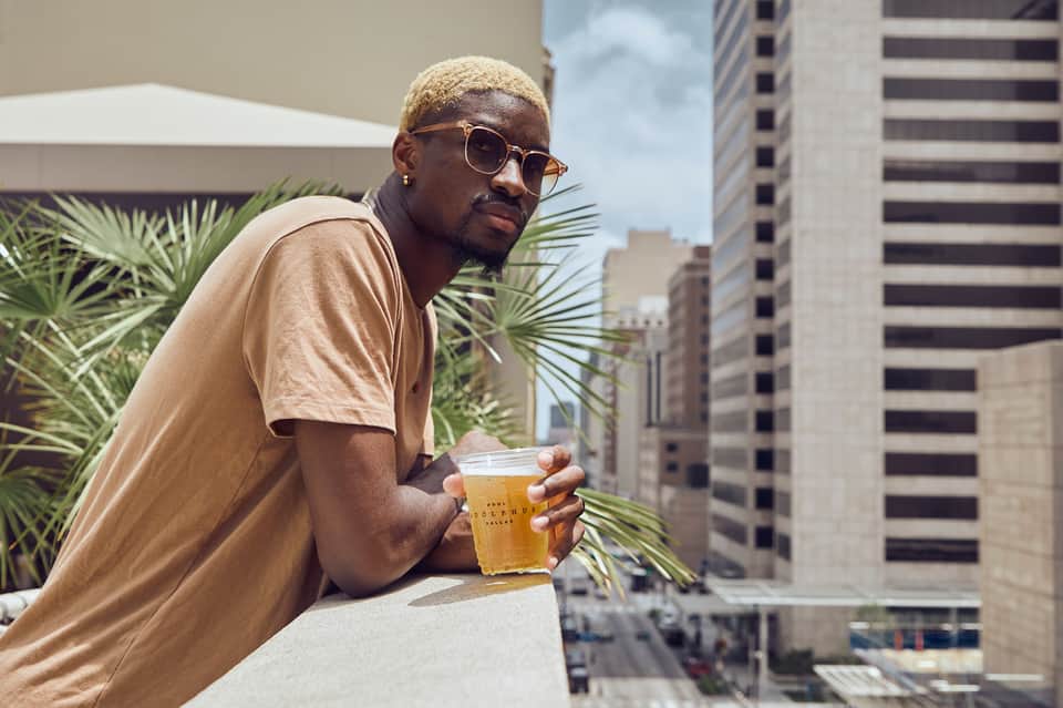 Man in beige shirt and sunglasses holding a drink on a rooftop terrace overlooking city skyline