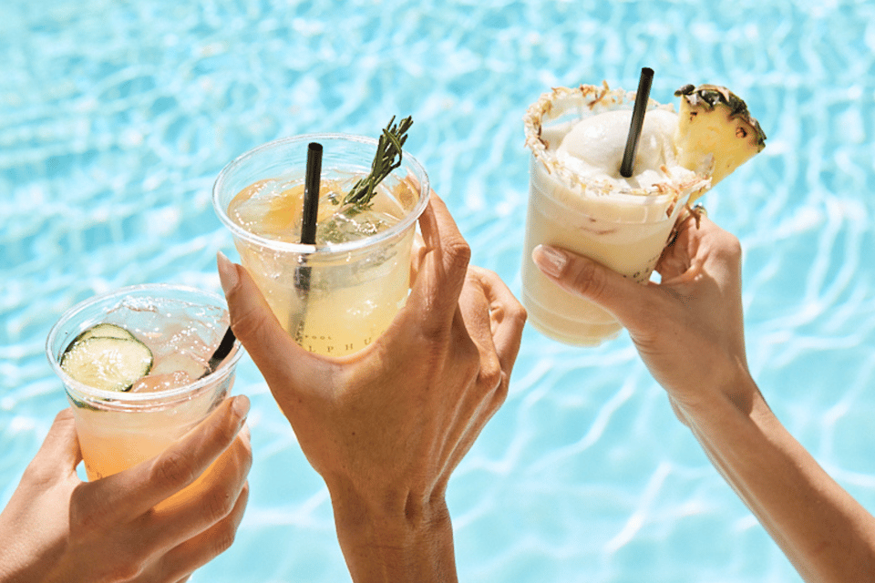 Three colorful cocktails held by hands over a sparkling blue pool with fruit garnishes and black straws
