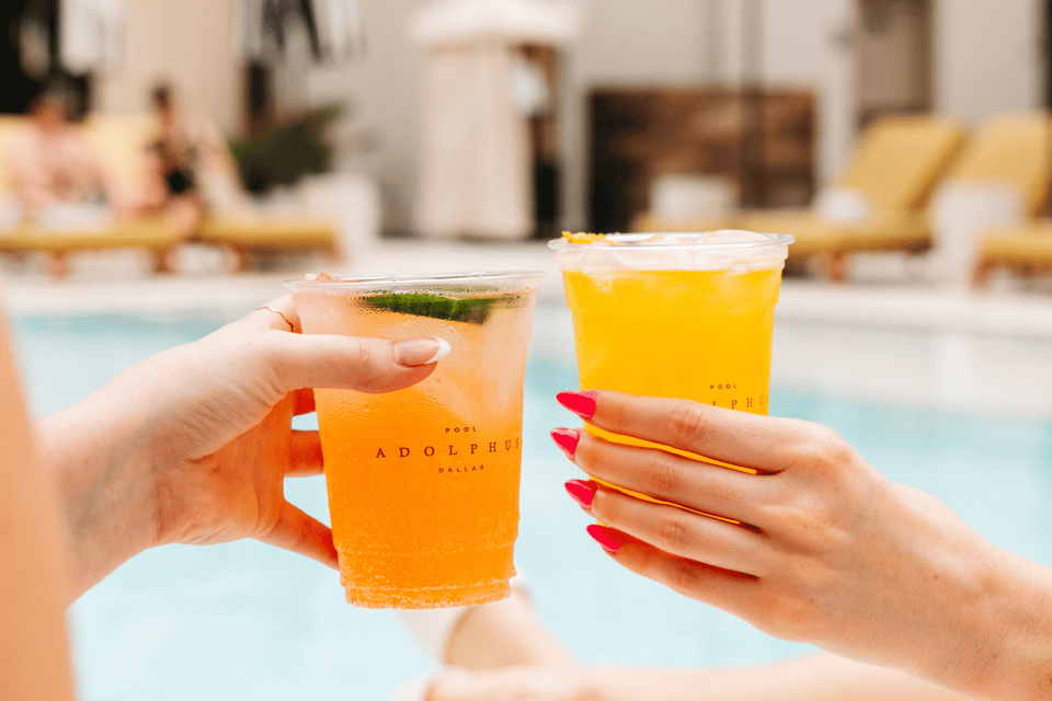 Two colorful cocktails held by hands with red nails by a pool at Adolphus Dallas hotel