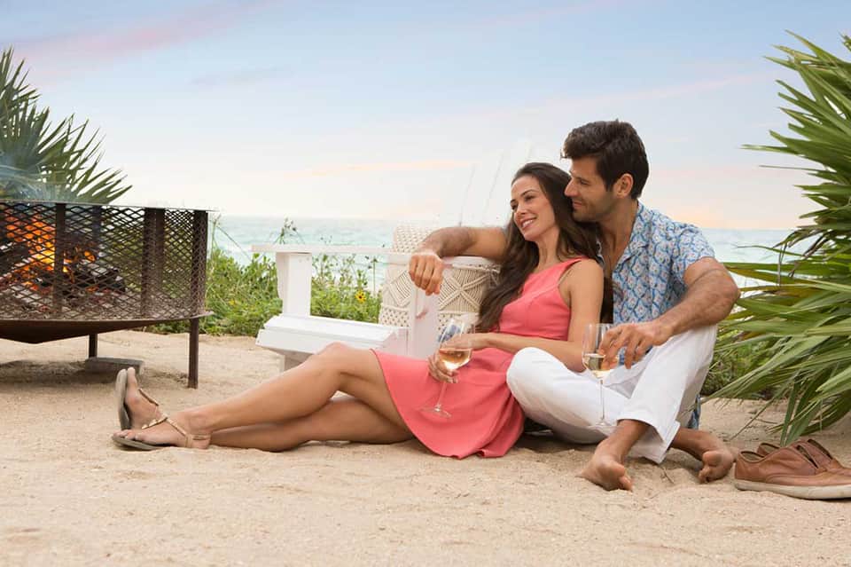 Couple in casual attire enjoying wine on a sandy beach with ocean view at sunset