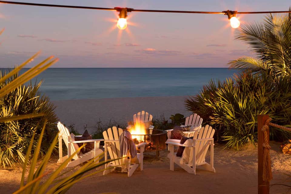 Beachfront fire pit with white Adirondack chairs at dusk, lit by string lights and palm trees