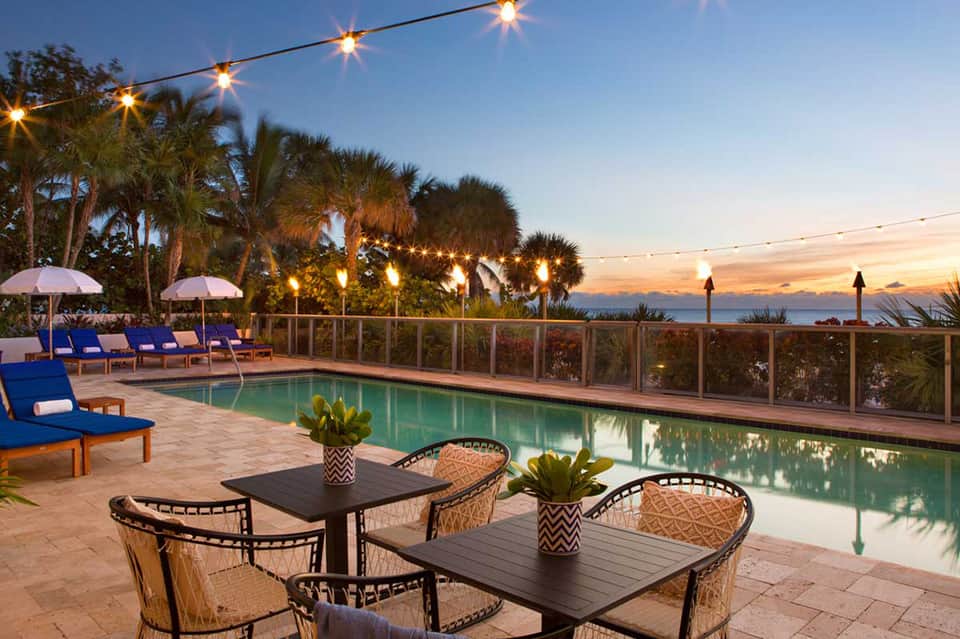 Twilight poolside terrace with lounge chairs, string lights, palm trees, and ocean view