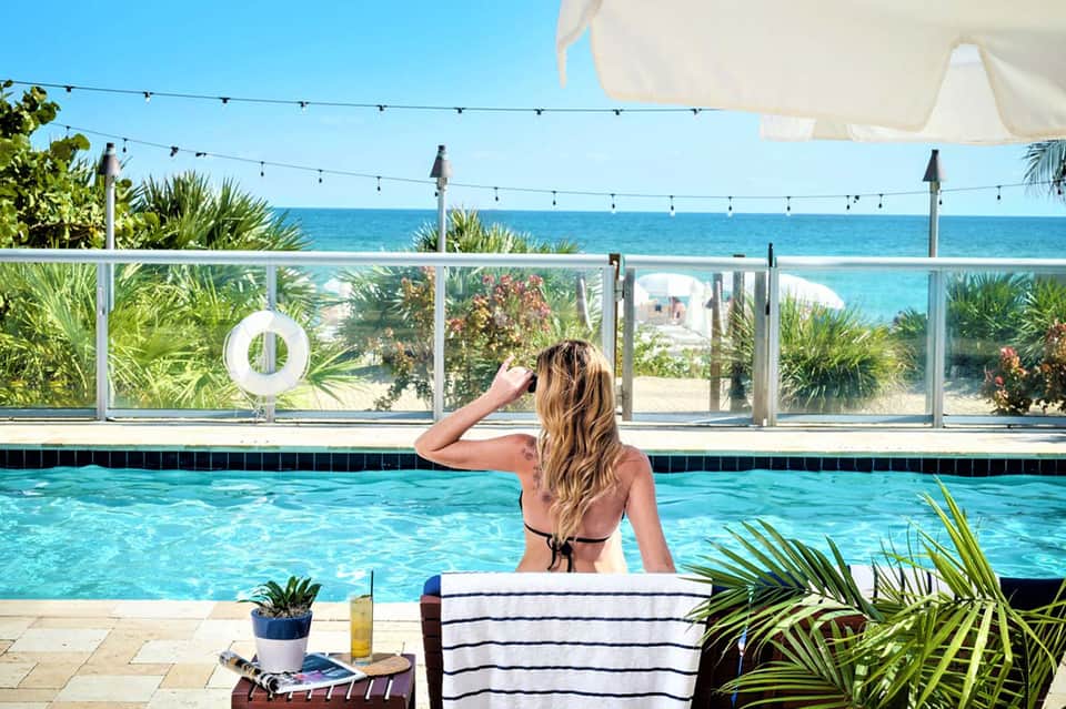 Woman in bikini relaxing by outdoor pool overlooking ocean with palm trees and beach cabanas