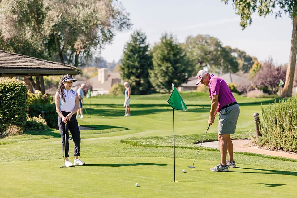Golfers on manicured fairway with flag pole, trees, and clubhouse shelter on sunny day