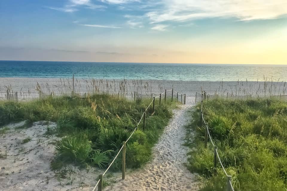 Sandy beach pathway with dune grass, wooden railings, and calm ocean at sunset