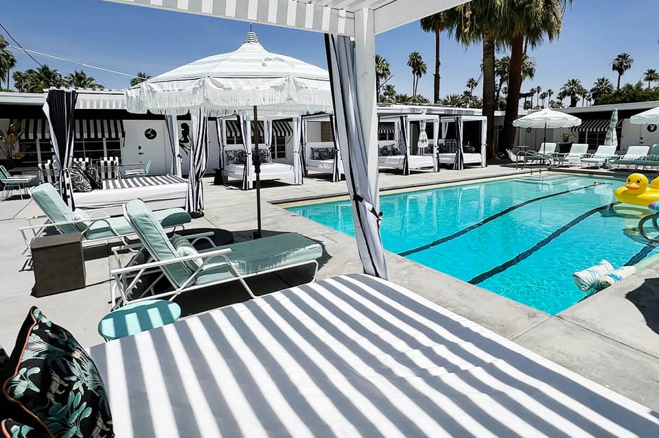 Resort pool with striped lounge chairs, white umbrellas, and palm trees under clear blue sky