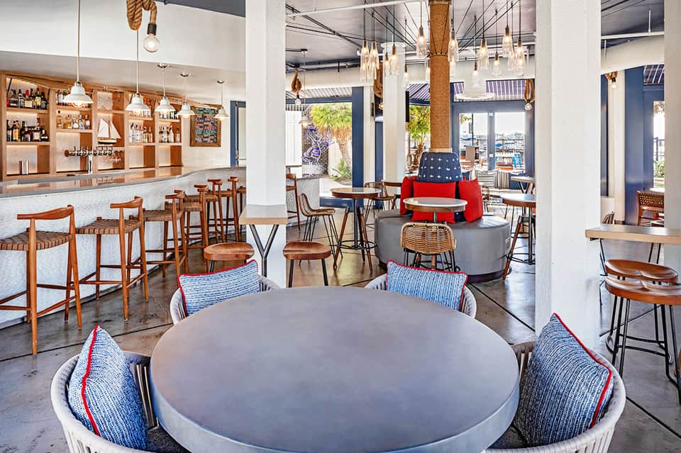 Modern hotel common area with bar, wooden furniture, round tables, and ocean views through large windows
