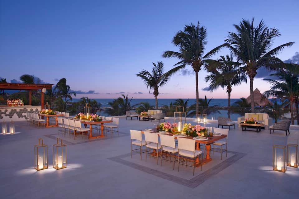 Elegant beachfront event space at dusk with palm trees, ocean view, and white dining furniture