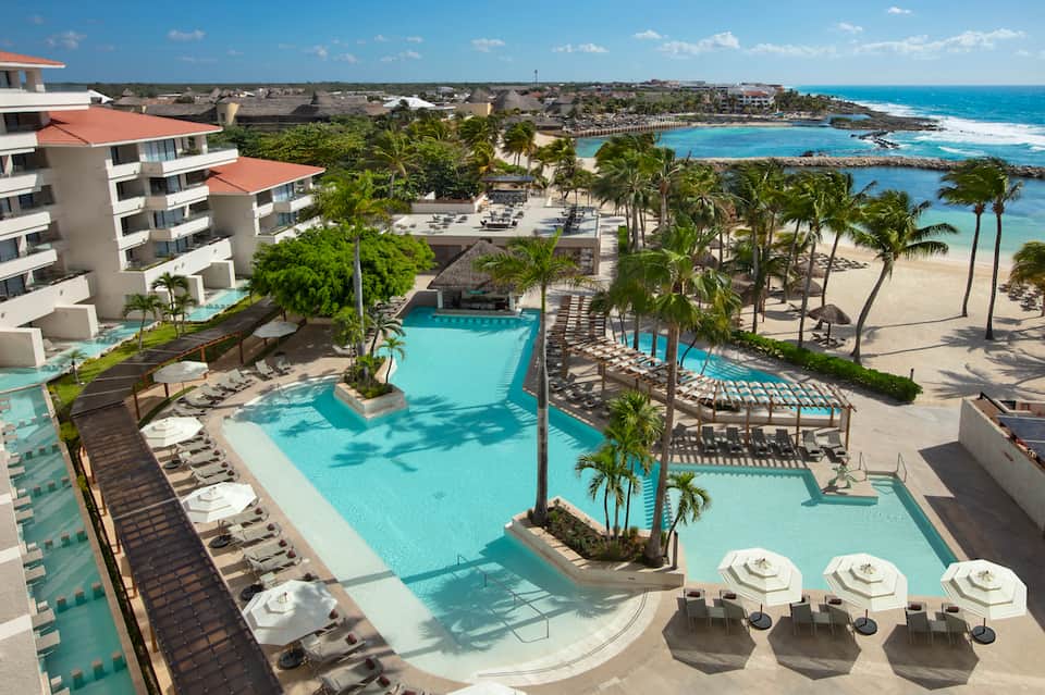 Resort pool complex with turquoise water, palm trees, beach access, and modern white building