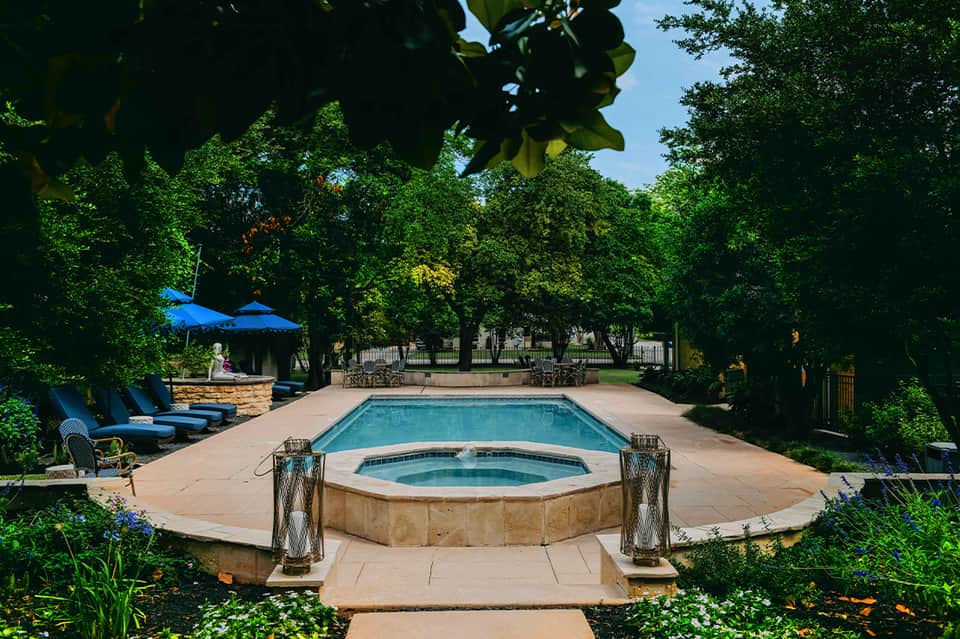 Resort pool with hot tub, loungers, blue umbrellas, and lush trees surrounding patio area