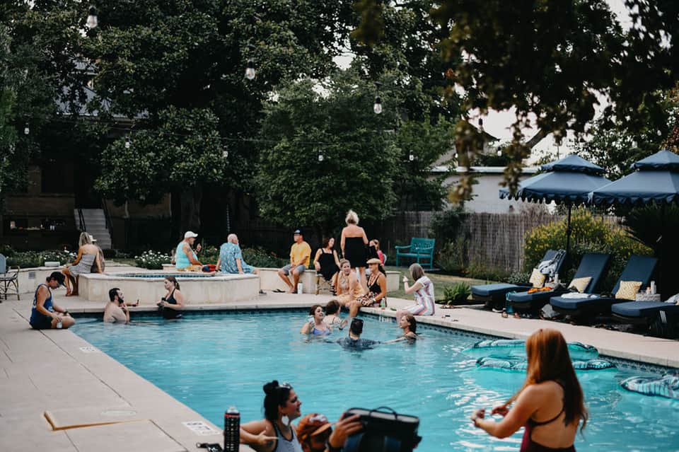 Busy resort pool with guests swimming and relaxing on lounge chairs surrounded by shade umbrellas and mature trees