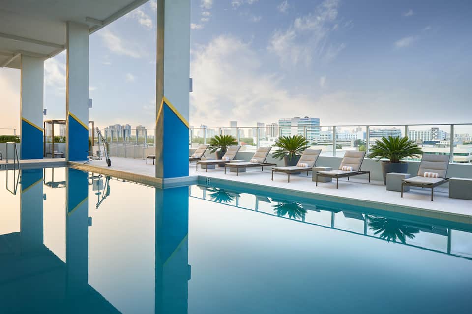 Indoor lap pool with city skyline views, lounge chairs, and palm plants on terrace