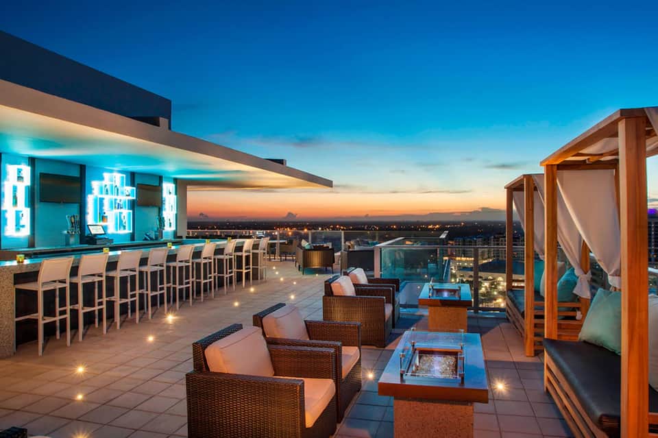 Rooftop bar at dusk with turquoise lighting, seating, and panoramic city views