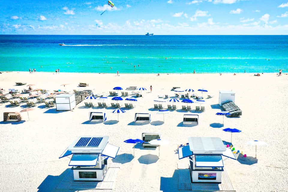 Pristine beach with turquoise water, white sand, blue umbrellas, lounge chairs, and beachgoers swimming