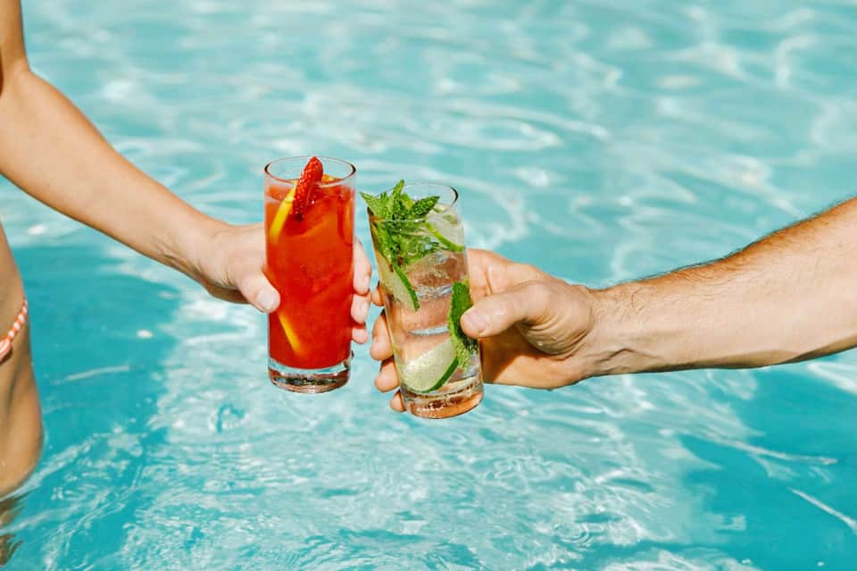 Two colorful cocktails held by hands over turquoise swimming pool water