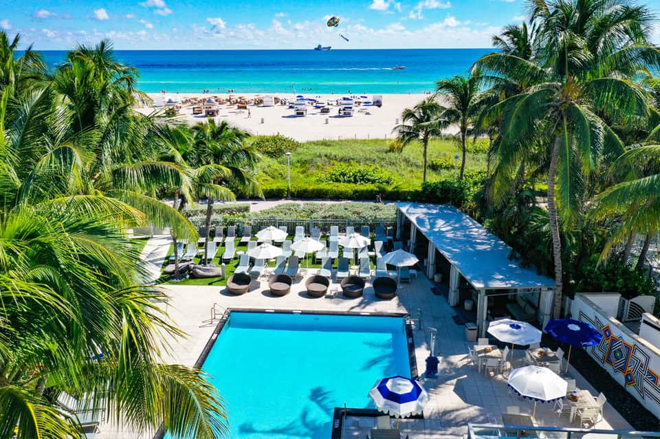 Aerial view of beachfront resort with pool deck, palm trees, and sandy beach with umbrellas
