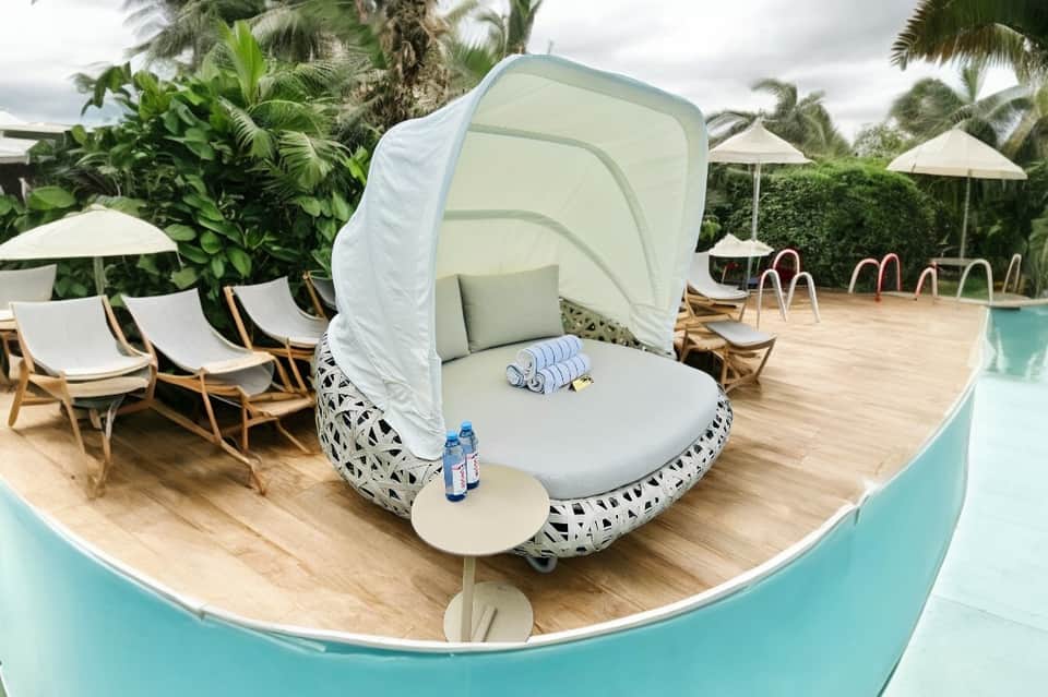 Resort poolside with white canopy daybed, lounge chairs, and palm trees under cloudy sky