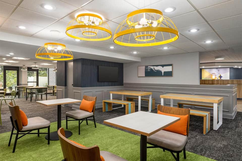 Modern hotel common area with yellow pendant lights, dining tables, and orange accent chairs