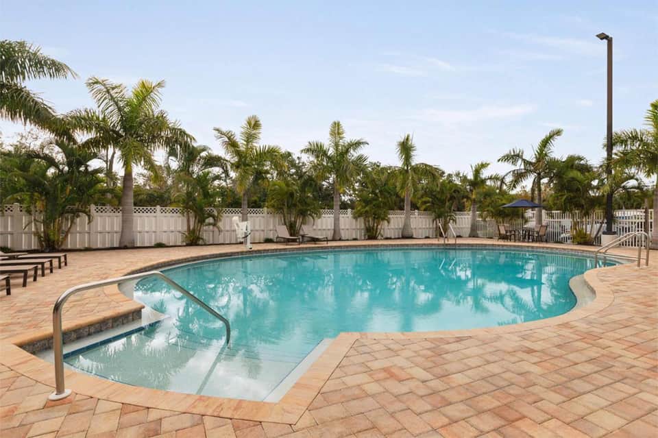 Resort pool surrounded by palm trees, white fence, and lounge chairs under clear sky