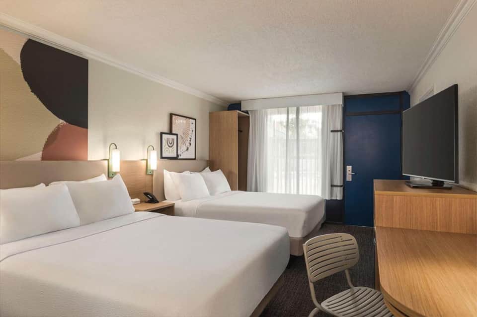 Modern hotel room with two queen beds, wood furnishings, blue accent wall, and waterfront views