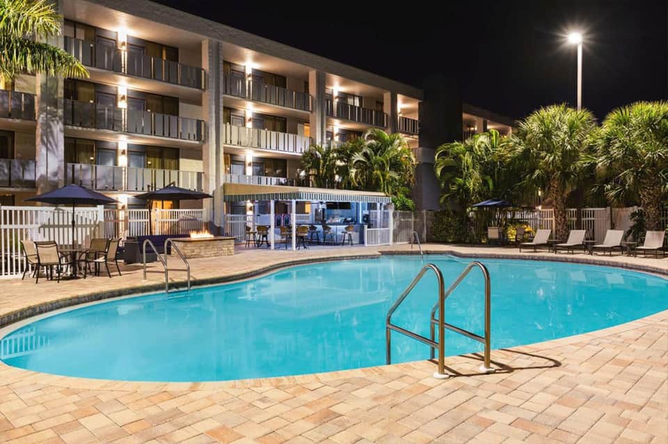 Illuminated resort pool at night with multi-story hotel building, palm trees, and lounge seating area